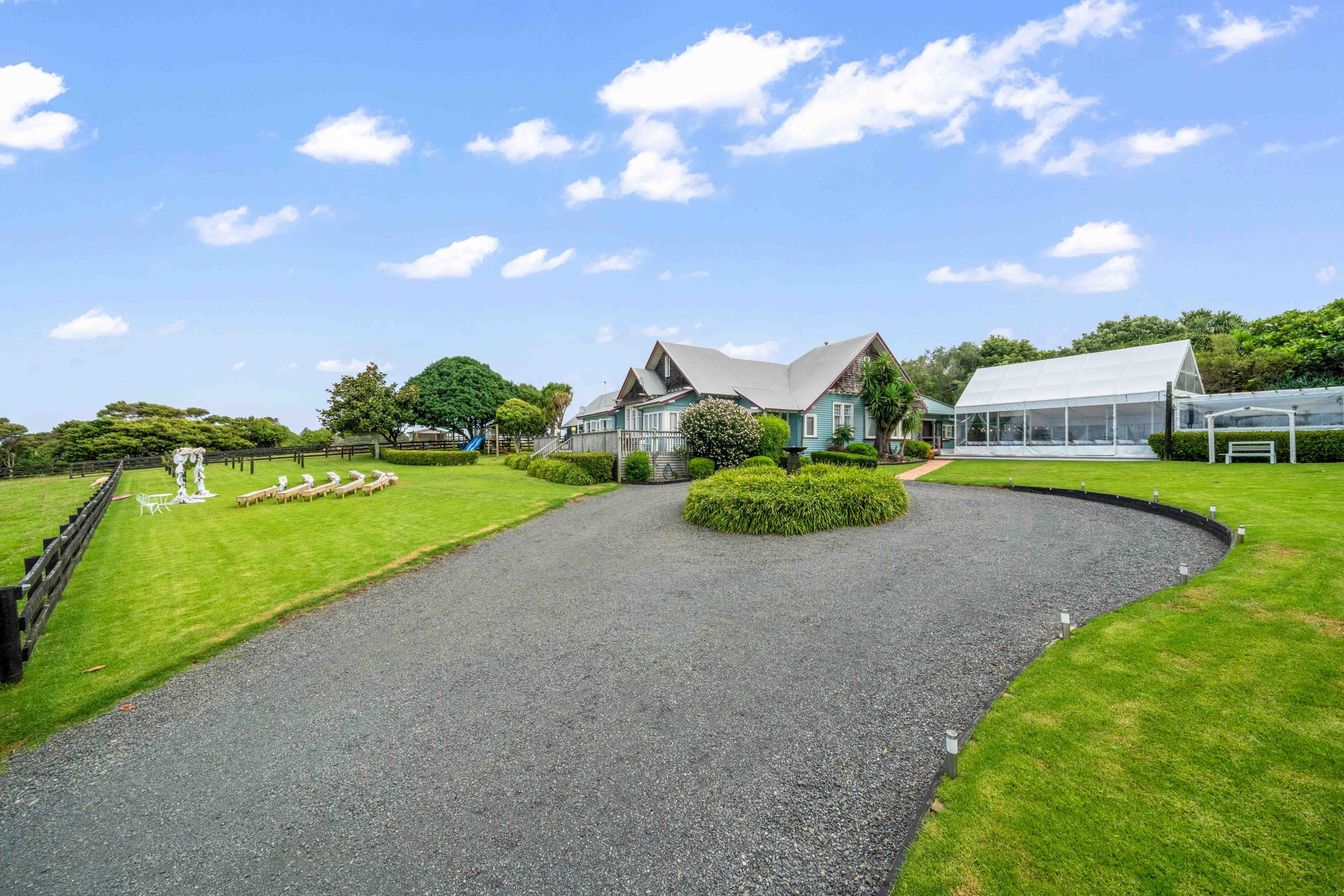 Gravel driveway curving to a dark-trimmed country house with steeply pitched roof, large lawn, white marquee to one side