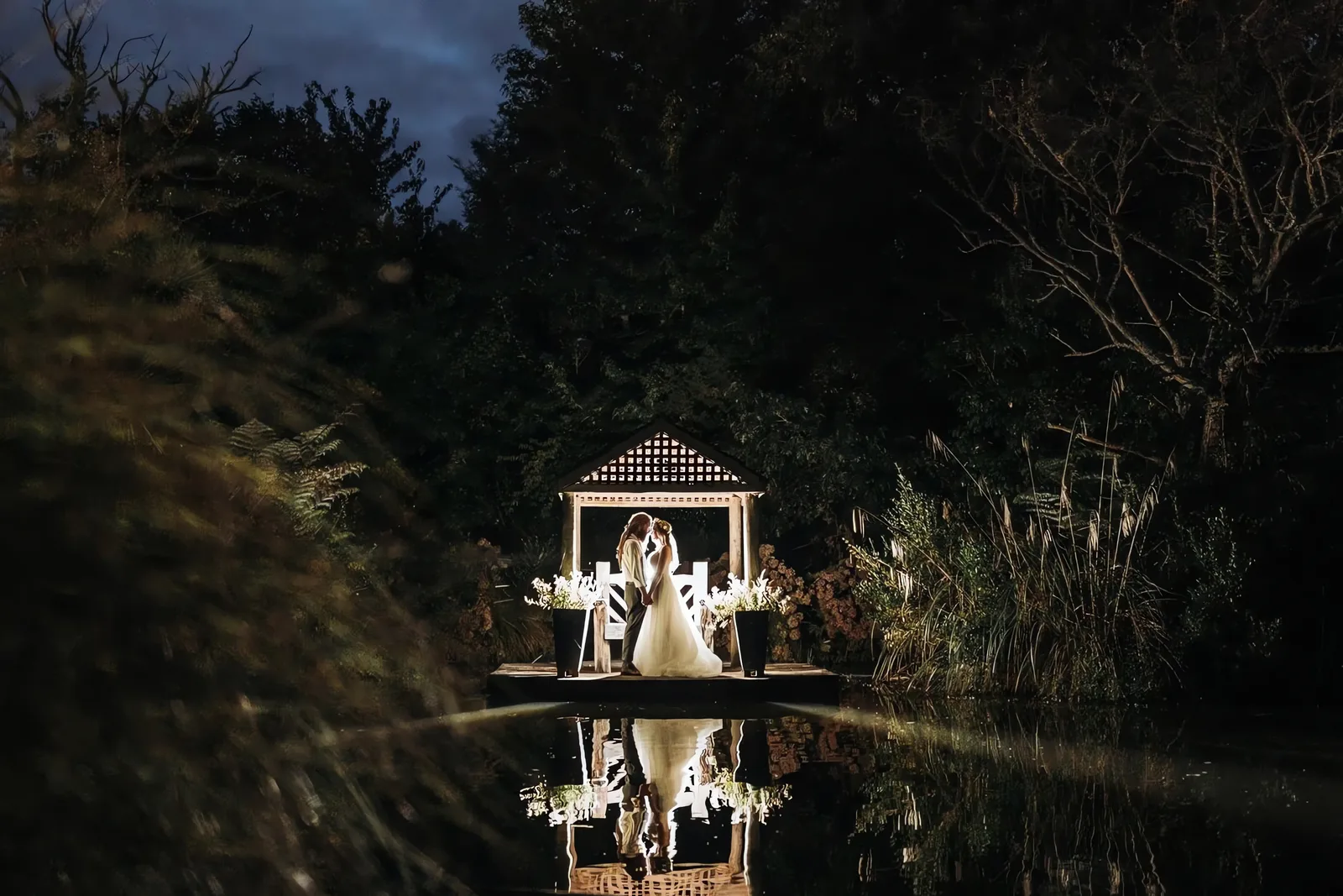 Illuminated gazebo on a jetty reflected in a still pond at night, surrounded by trees