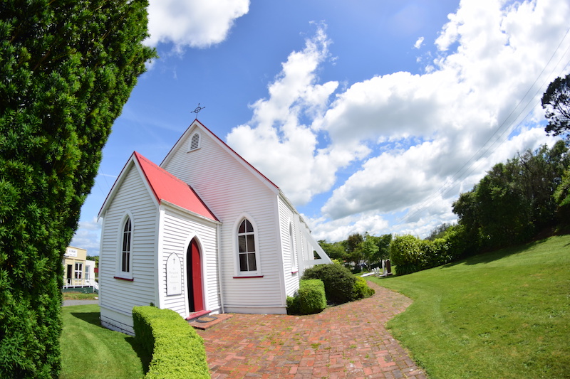 Small white-painted timber church with red-trimmed gable and red door, brick path entrance
