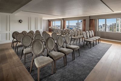 Neutral-toned meeting room with rows of upholstered chairs, two large windows, pale timber flooring