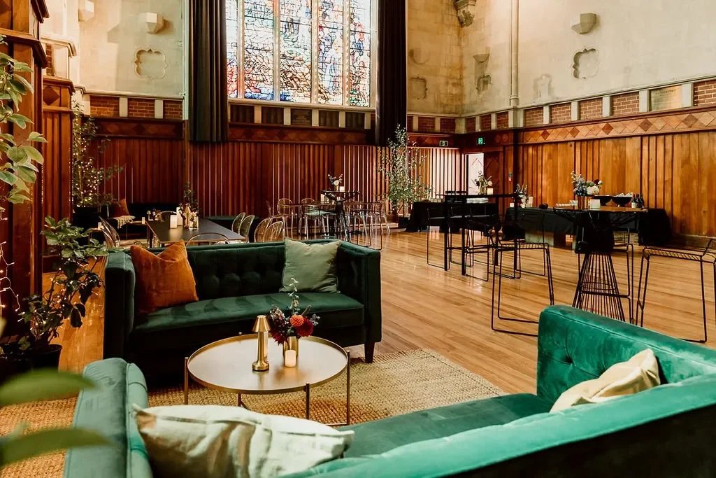 Church hall interior with dark timber panelling, stained-glass windows, and velvet sofas