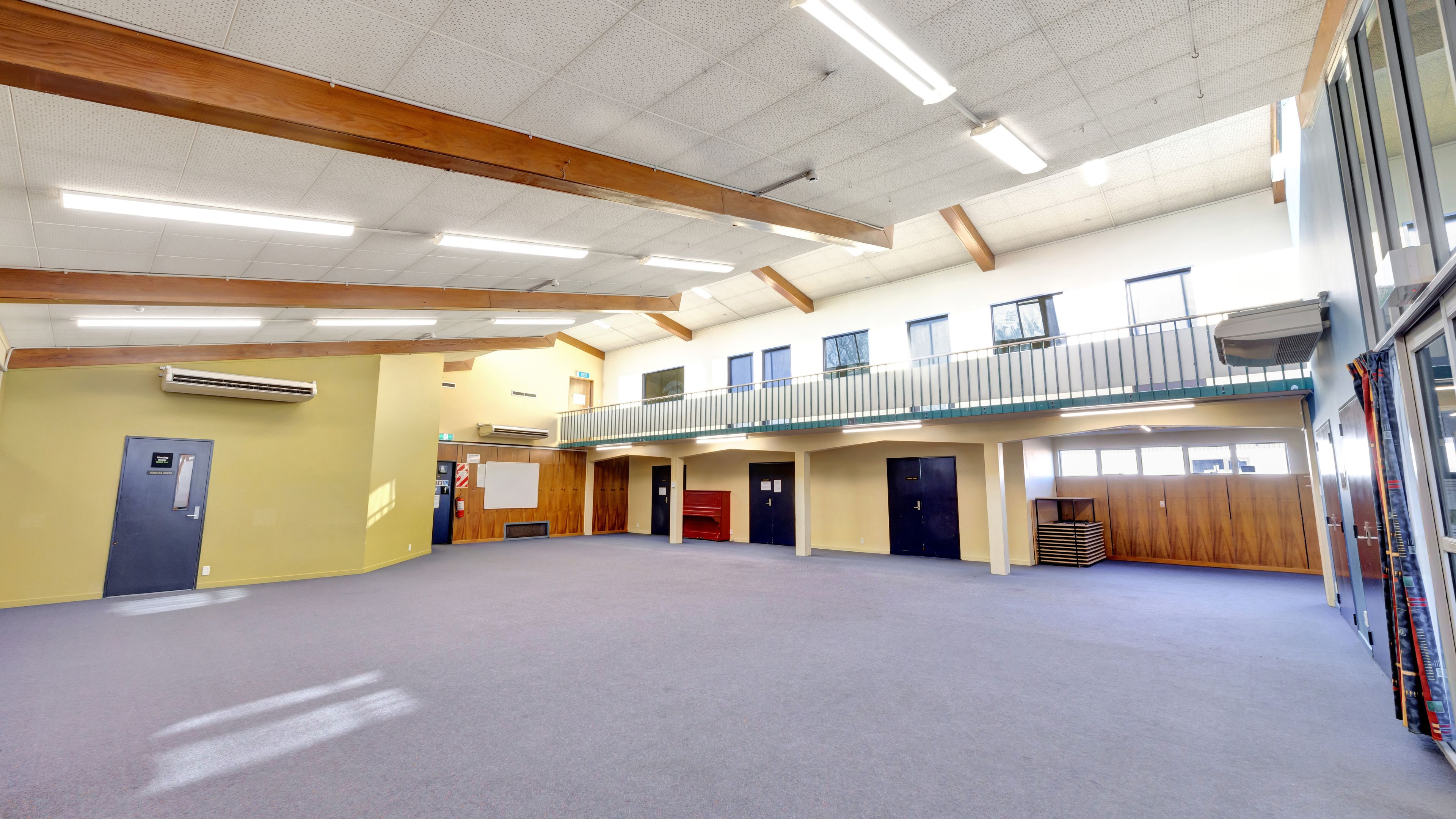 Large cleared hall with grey carpet, exposed timber roof beams, mezzanine balcony, and high windows