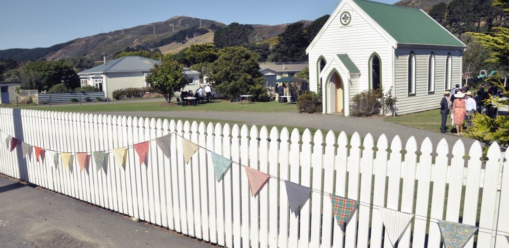 Small white timber chapel with green roof and arched entry, beside a white picket fence with bunting