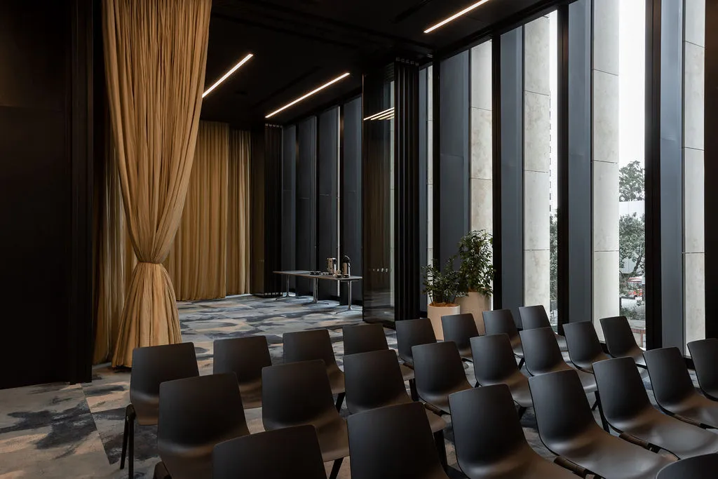 Dark-toned meeting room with floor-to-ceiling windows, city views, rows of dark chairs and gold drape partition