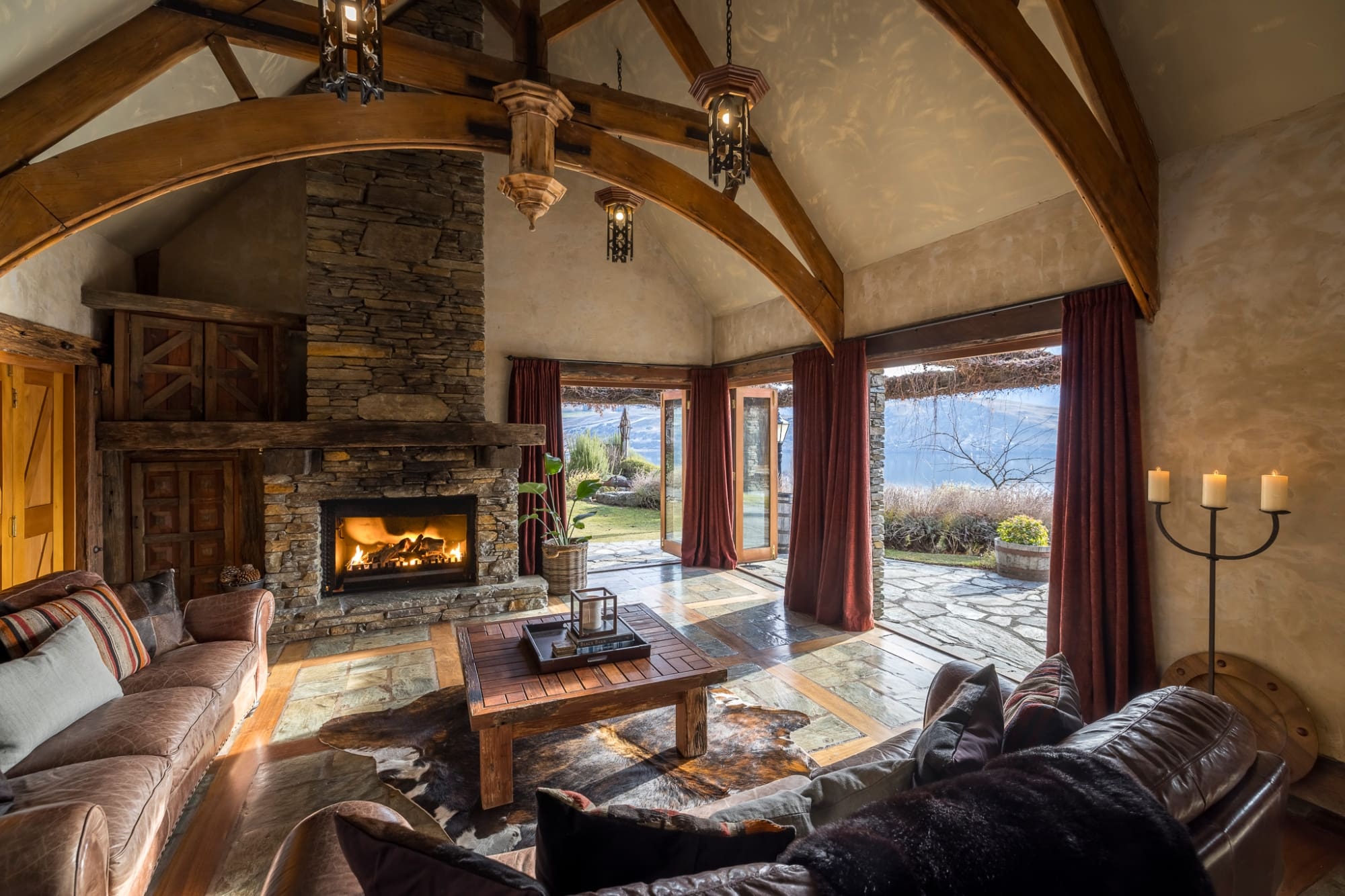 Stone fireplace lounge with vaulted timber beams, leather sofas, and open doors to garden view