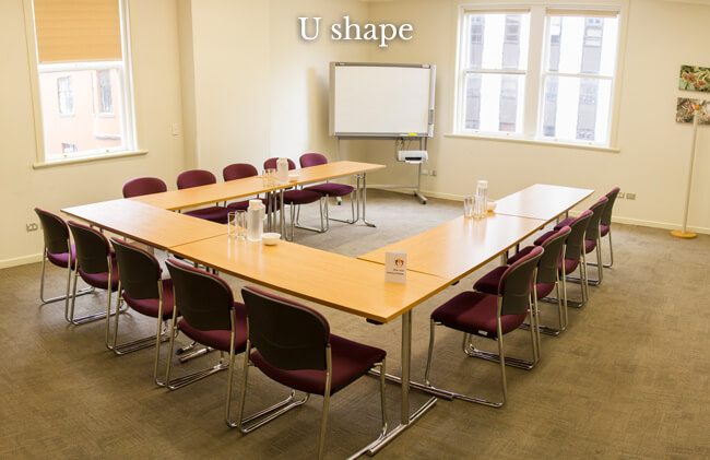 Carpeted meeting room with U-shaped timber tables, purple chairs, and whiteboard by windows