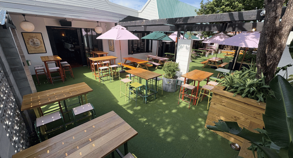 Open-air courtyard with timber picnic tables, coloured stools, market umbrellas, and turf underfoot