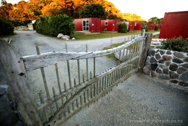 Rustic wooden entrance gate onto a gravel driveway with red cabin buildings beyond at dusk