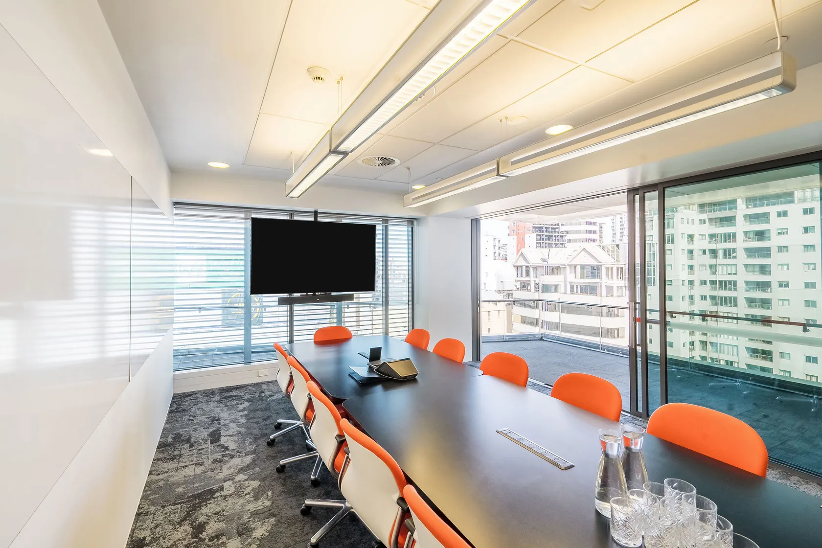 Long meeting room with dark table, orange chairs, wall-mounted screen, and city-view windows on two sides