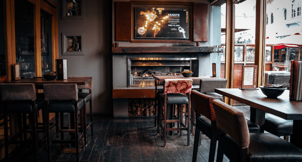 Dim bar room with open fireplace, upholstered high stools, dark timber tables, and street-facing windows