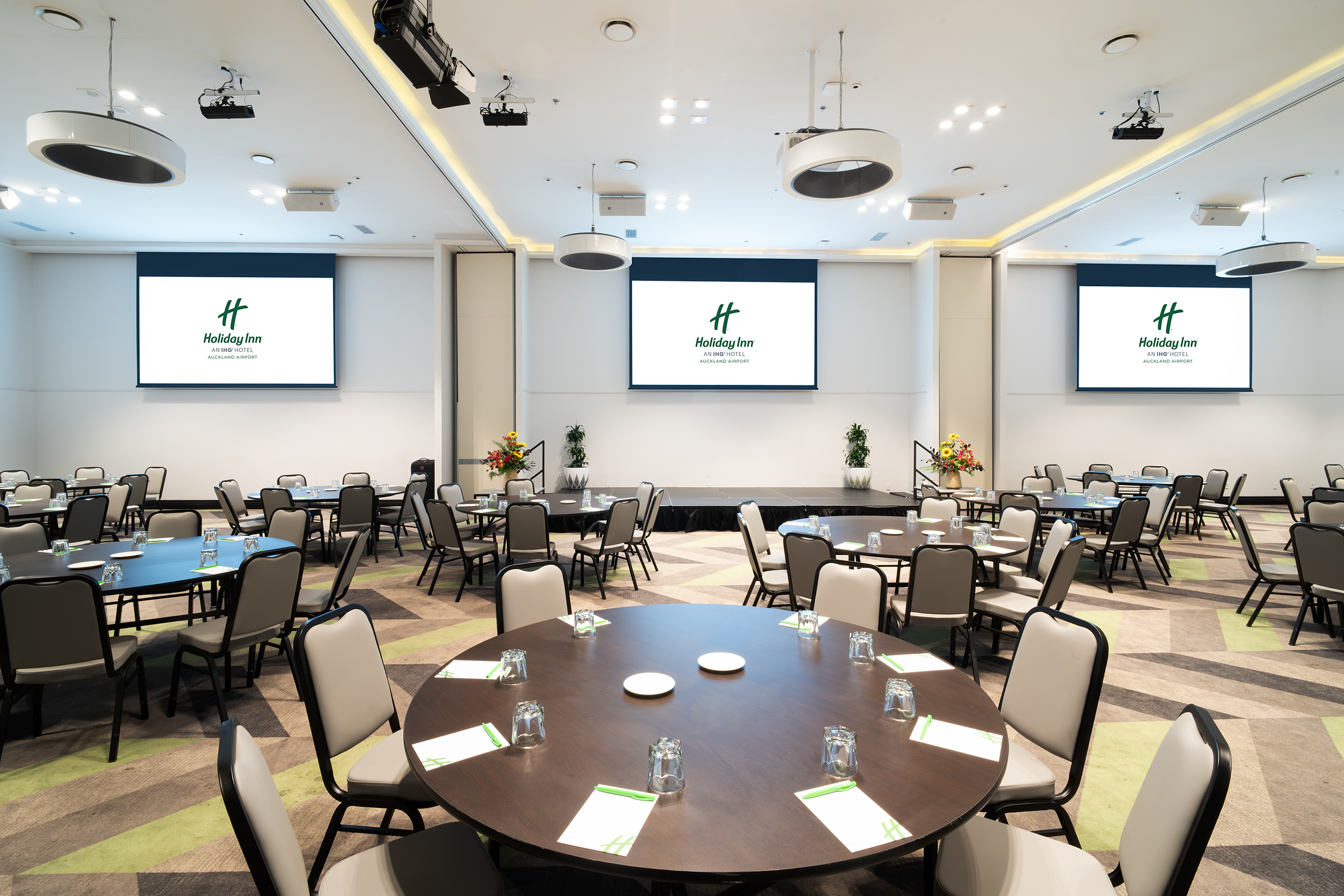 Large carpeted function room with round tables, three projection screens, and recessed ceiling lighting
