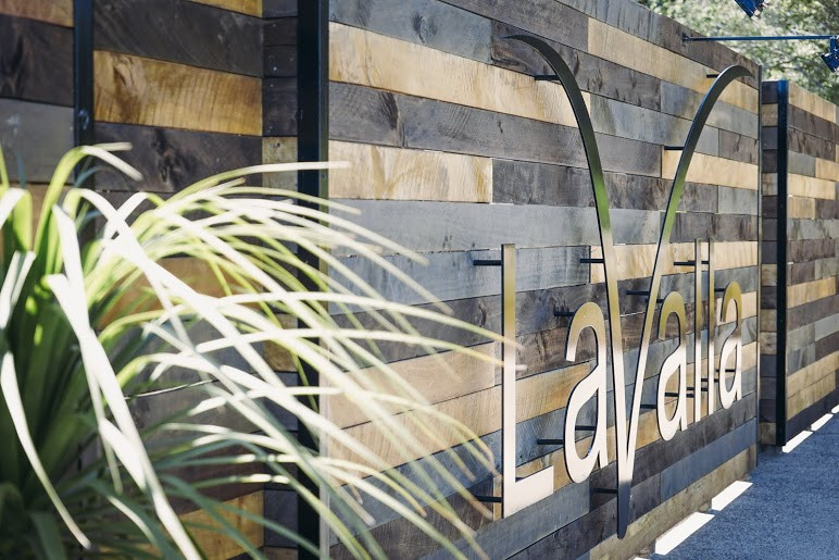 Horizontal timber-clad fence with a metal venue sign, green foliage in the foreground