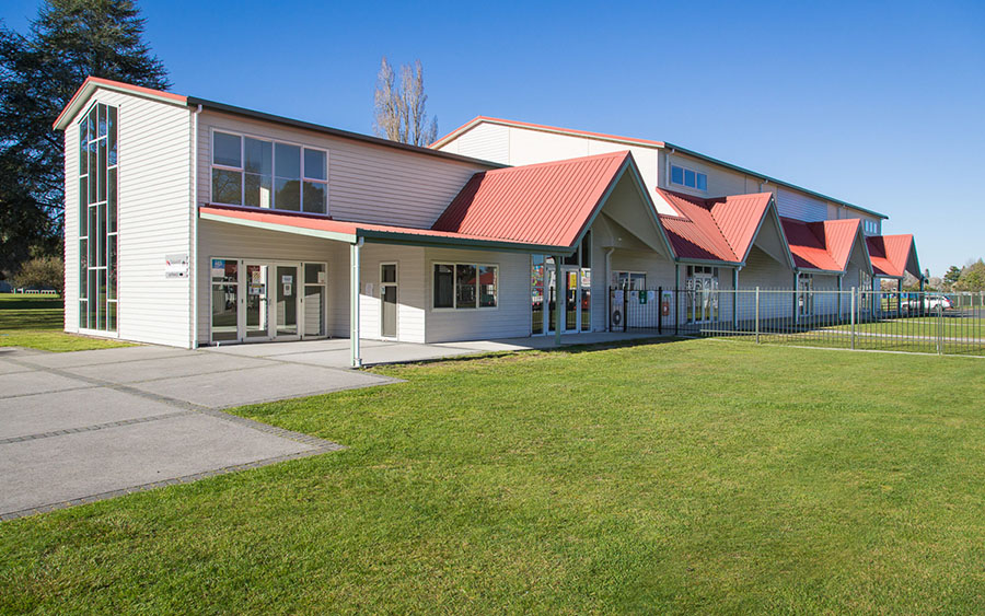 Long low building with white weatherboard cladding and red metal roof, set on a large lawn