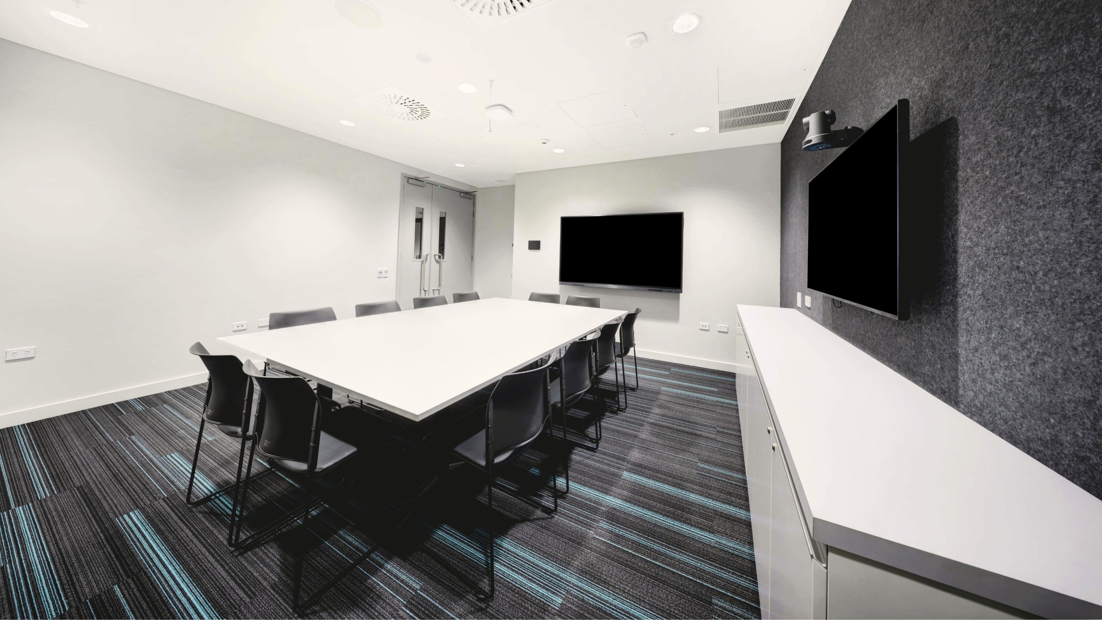 White meeting room with long white table, black chairs, two wall-mounted screens and patterned carpet