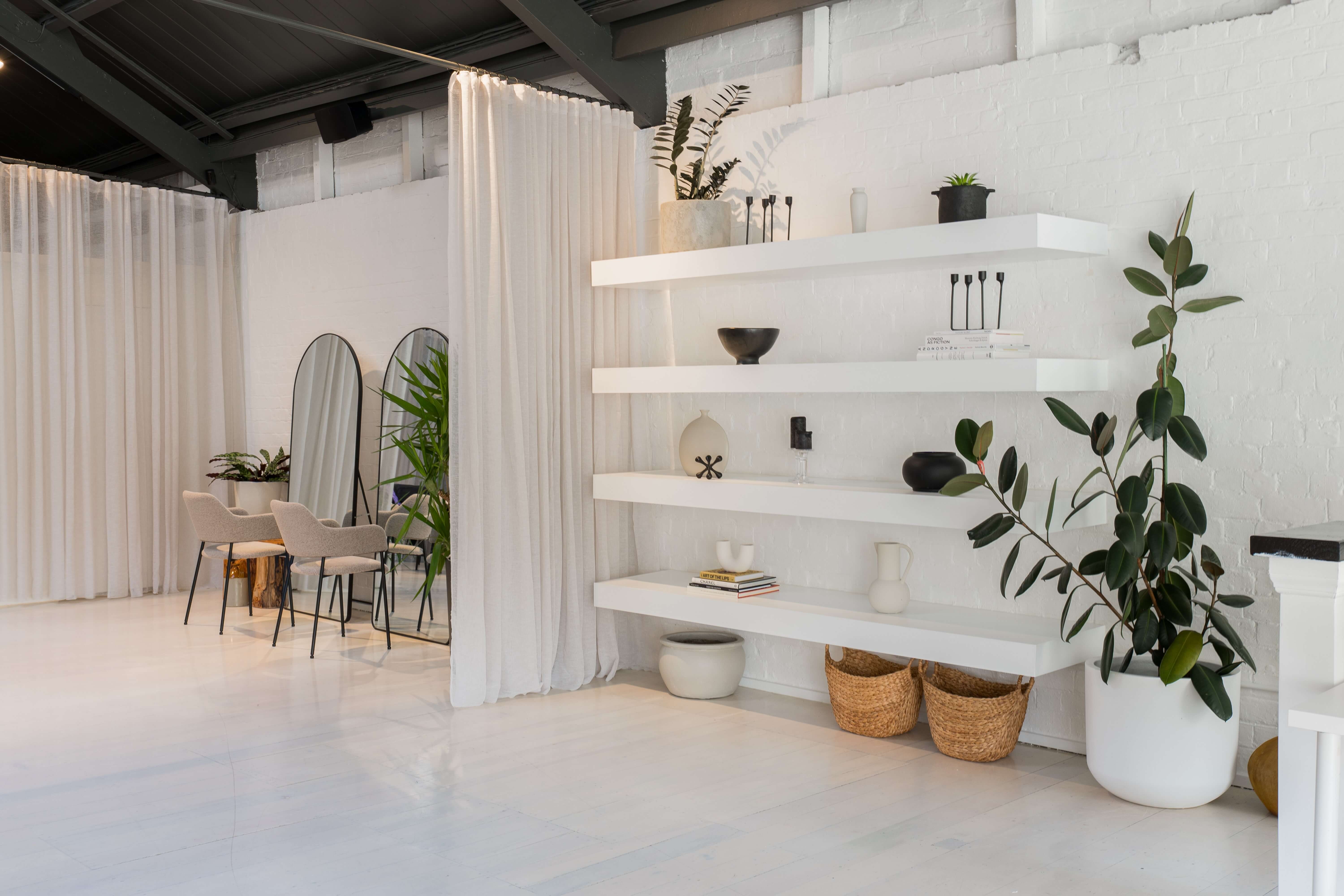 Bright white studio with open shelving, potted plants, sheer curtain partition, and arched mirrors