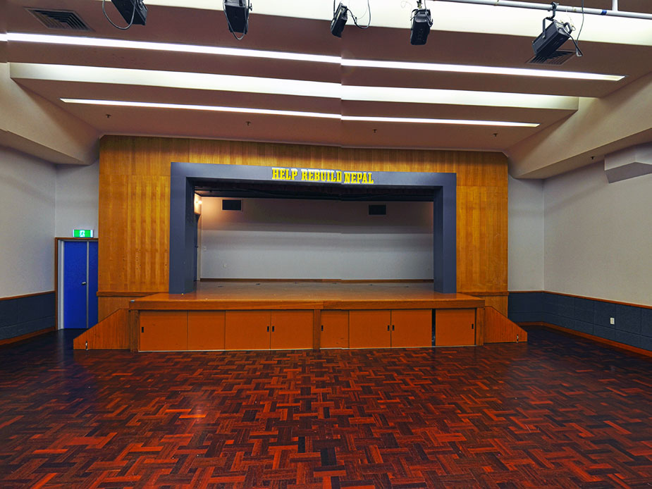 Hall interior with parquet floor, raised stage with proscenium arch, and stage lighting overhead