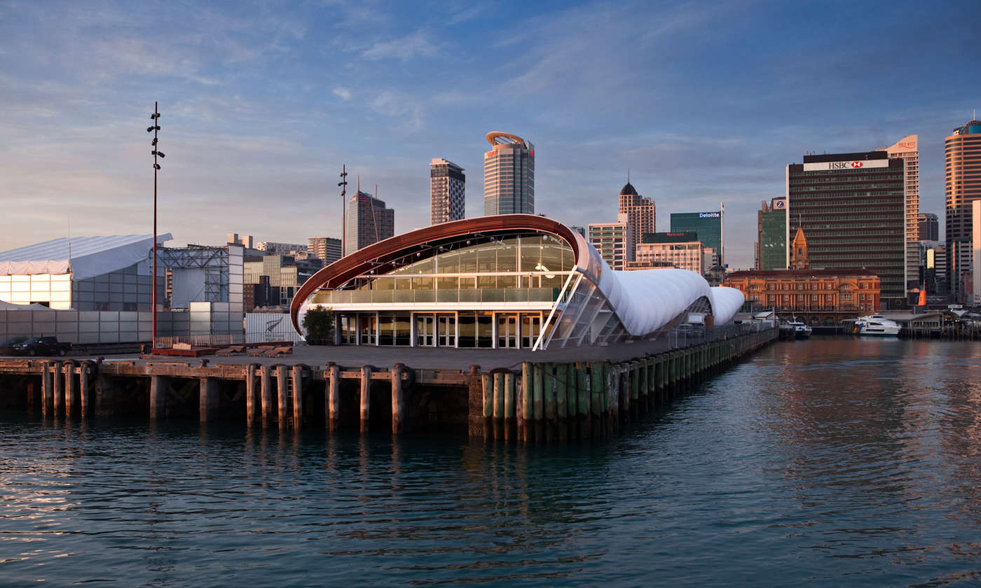 White barrel-vault pavilion on pier over water, Auckland city skyline at dusk behind
