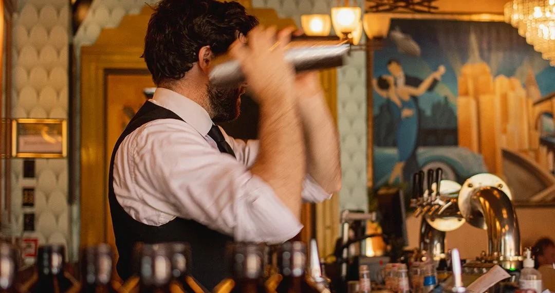 Bartender shaking cocktail behind bar with art deco mural and beer taps