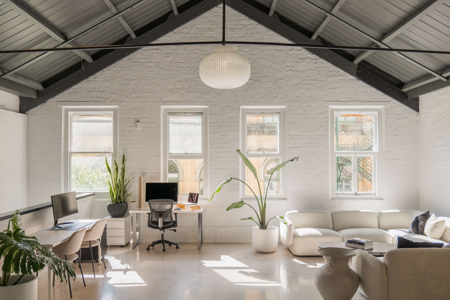 White-painted brick interior with vaulted steel-truss roof, lounge seating, desks, and tall plants