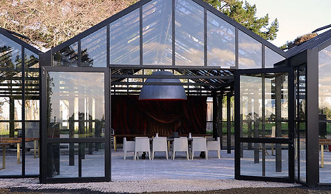 Black steel glasshouse interior with white chairs and dark hanging pendant, surrounded by trees