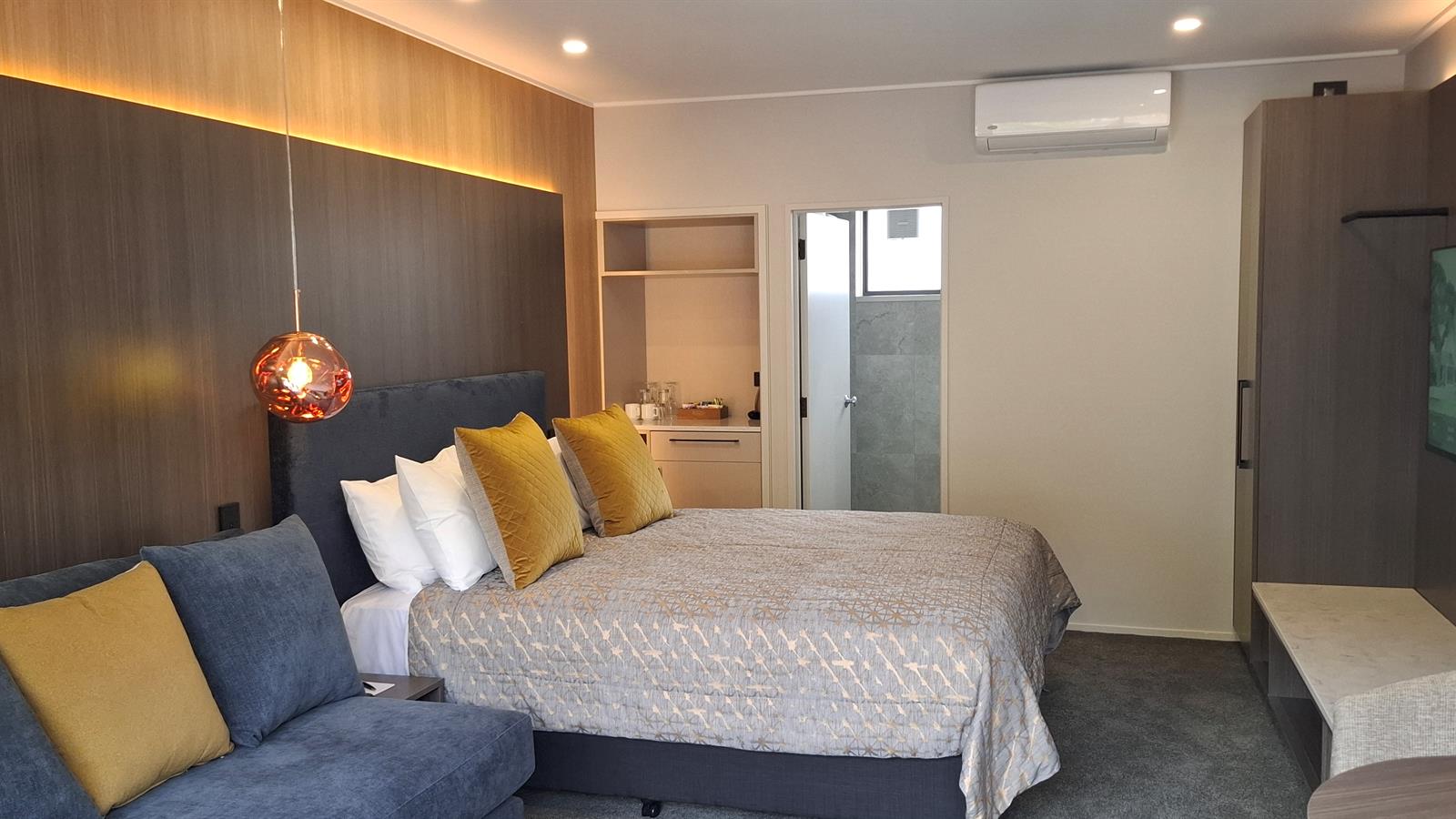 Hotel room with large double bed, yellow cushions, timber-panel feature wall, pendant light, and ensuite beyond