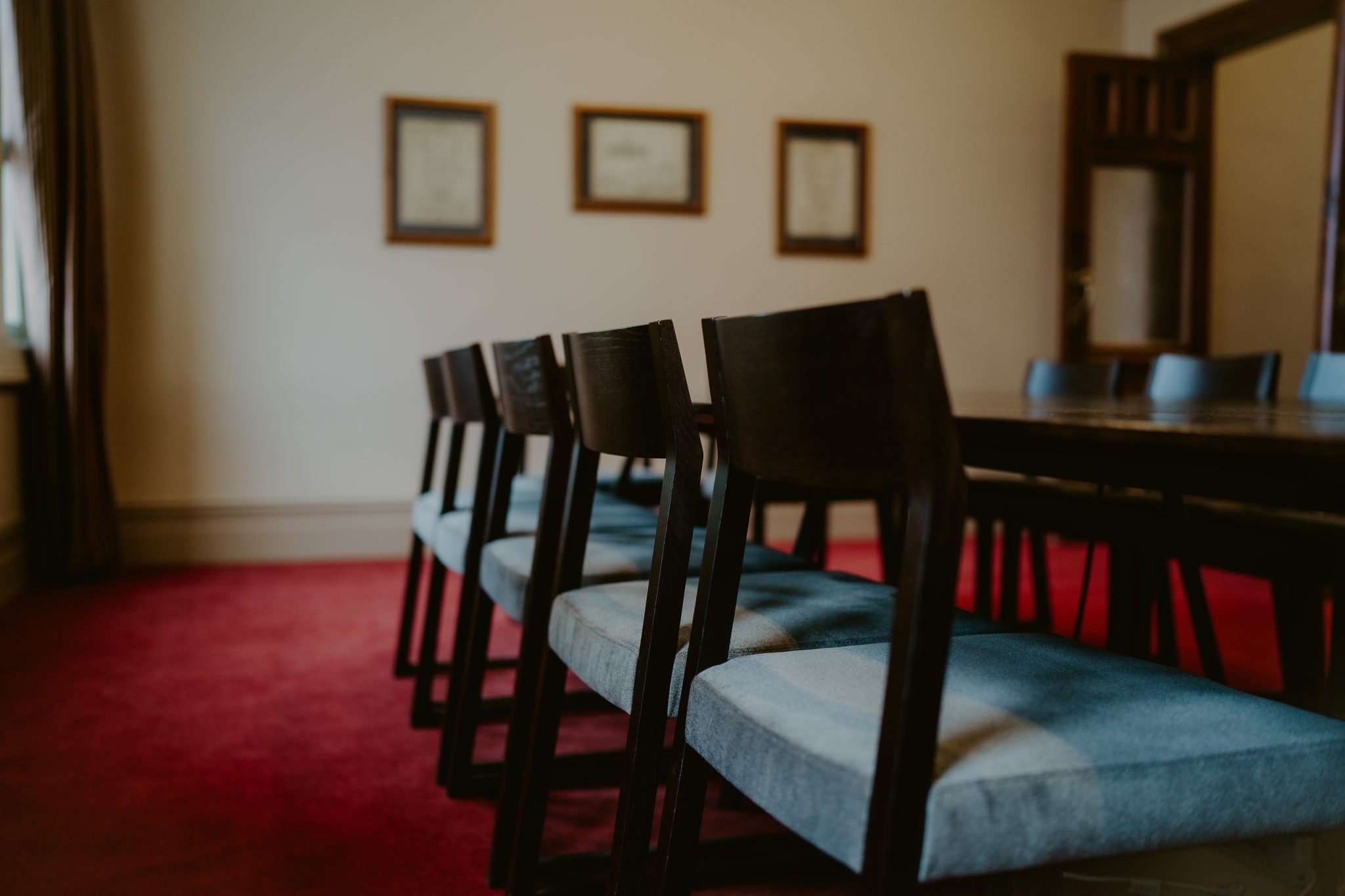 Dark-toned room with dark wood chairs and blue upholstered seats around a long table, framed prints on wall