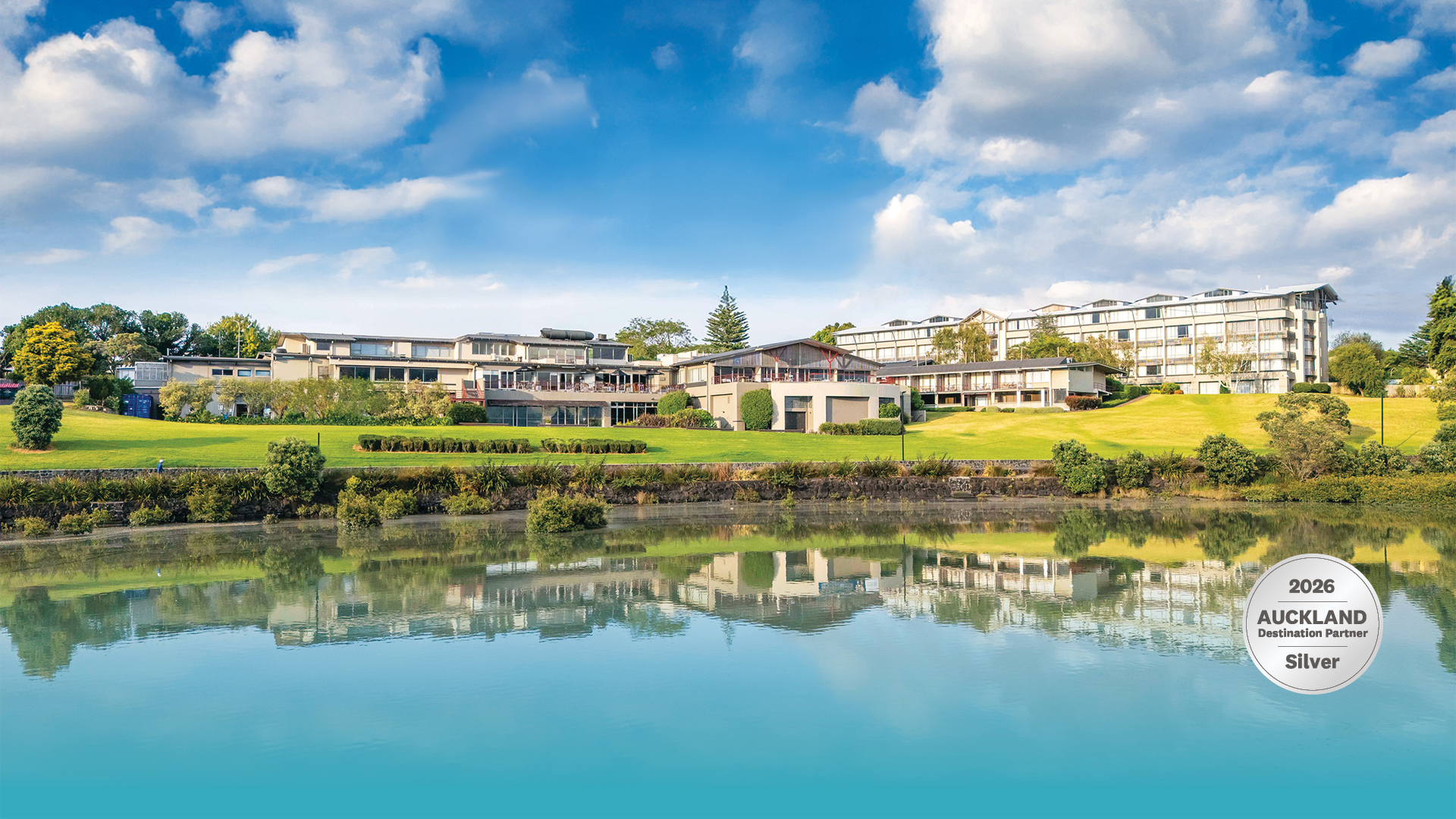 Multi-storey hotel complex reflected in still lake, surrounded by lawns and trees