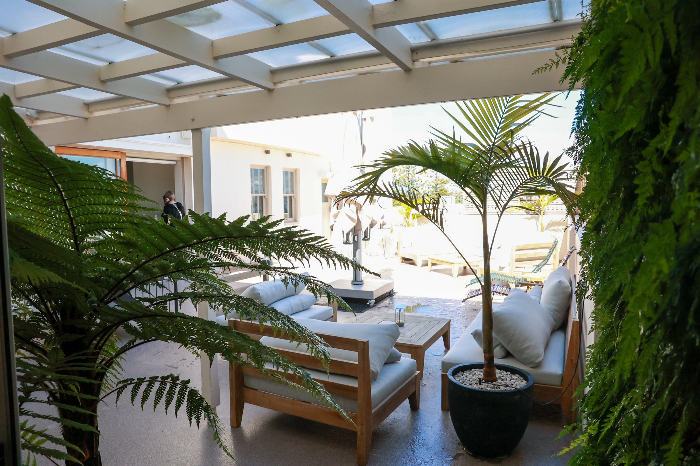 Covered outdoor terrace with white lounge furniture, potted palms, and fern wall, pergola roof