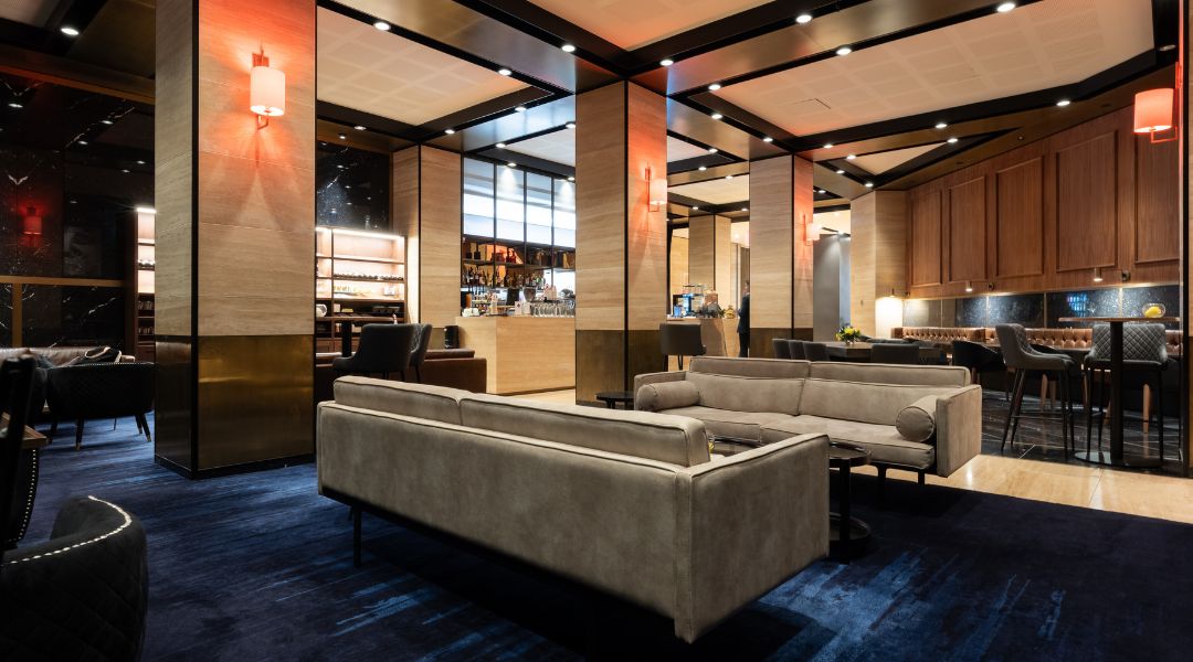 Lounge area with grey sofas, wood and mirror wall panels, warm ceiling lights, bar visible through opening