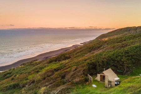 Canvas tent structure on grassed hillside at sunset, overlooking black-sand beach and ocean