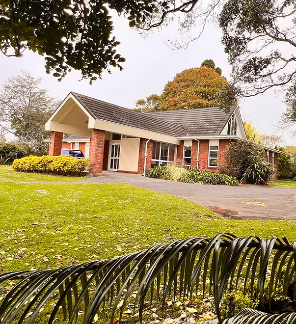 Low-set brick building with a pitched roof and covered entry, surrounded by lawn and trees