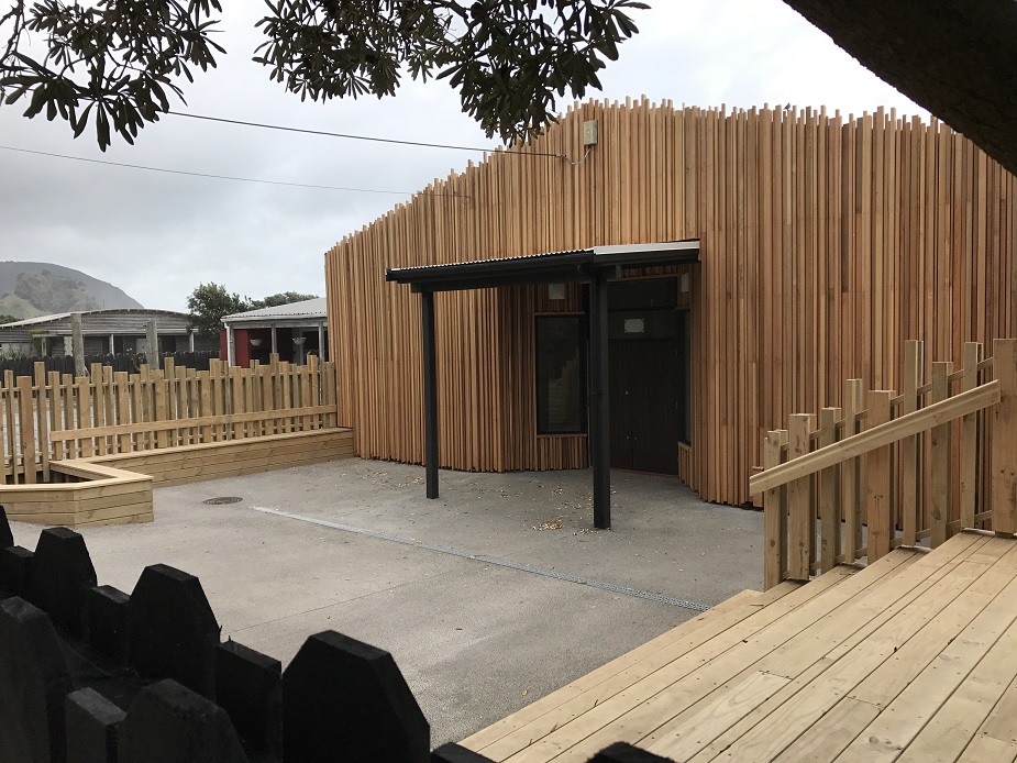 Rounded timber-battened building with covered entry porch, concrete forecourt and timber deck, hills behind