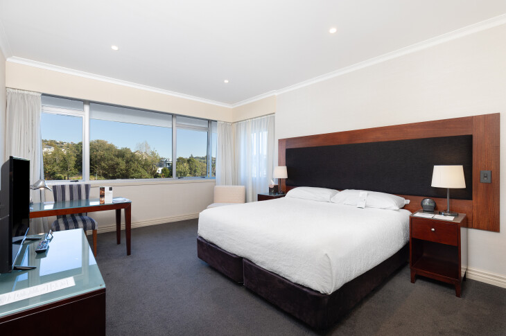 Hotel room with king bed, dark timber headboard, desk and windows to green view