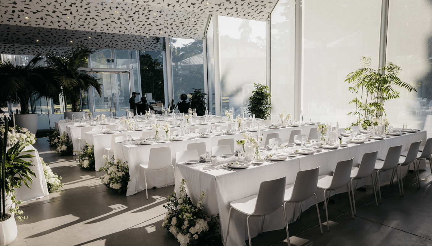 Glass-walled function room with long white tables and floral arrangements set for dining