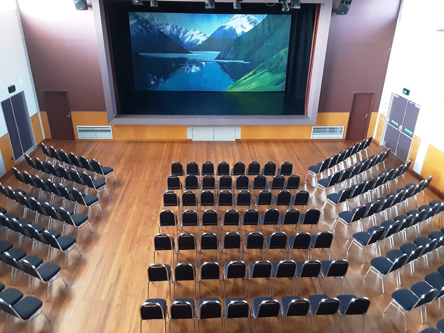 Auditorium with rows of black chairs facing a large projection screen on a raised platform, timber floor