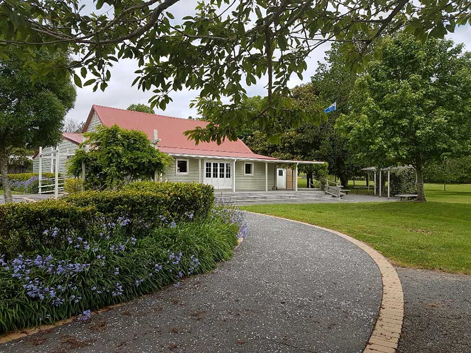 White weatherboard hall with red roof viewed along a curved driveway with flowering border