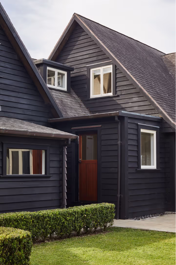 Dark-painted weatherboard homestead with steep gable roof, red timber door and clipped hedge border