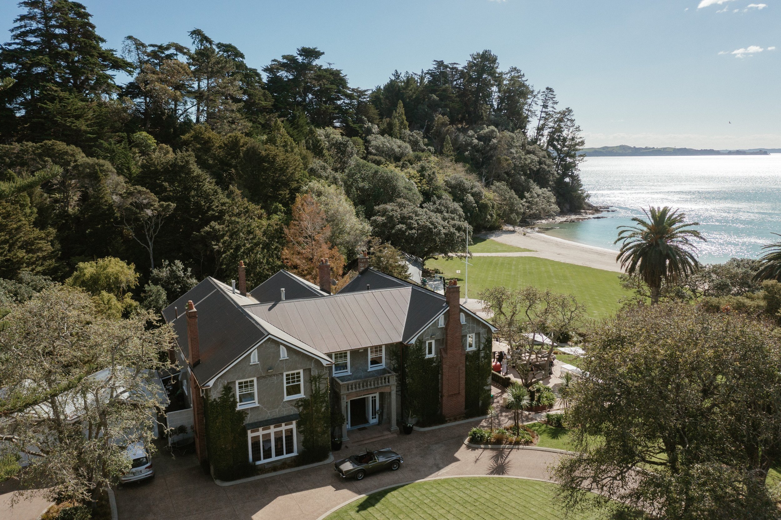 Aerial view of large historic stone manor with lawn, trees and beach frontage