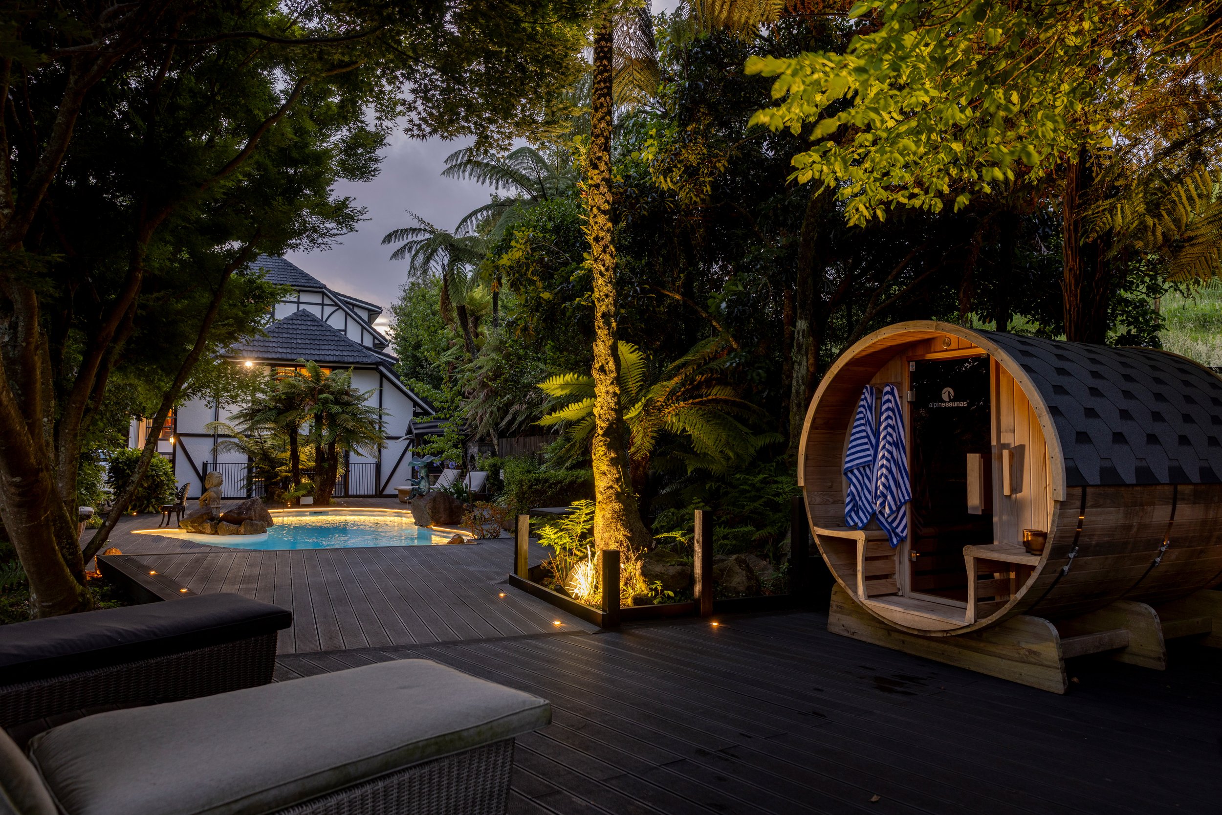 Deck area at night with barrel sauna, pool, and trees lit by ground lights