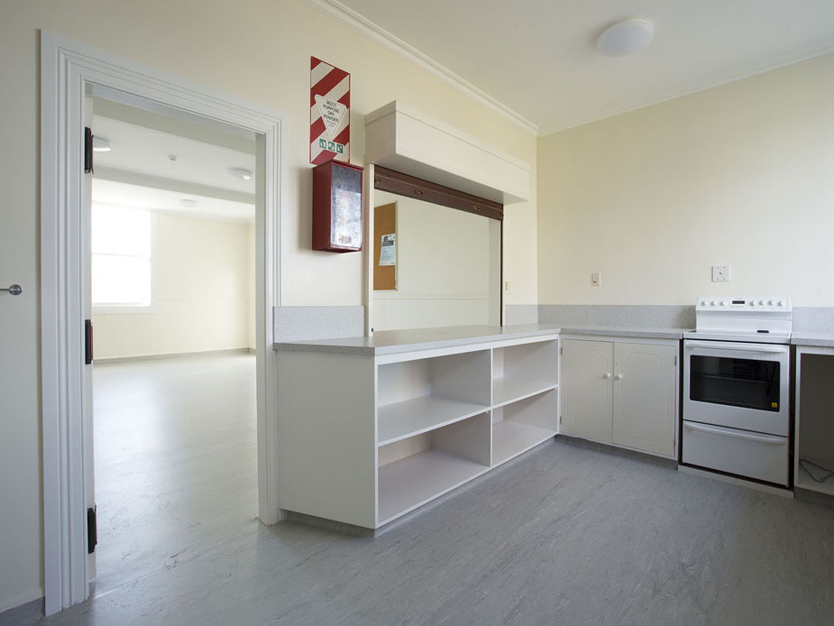 Cream kitchen with a serving counter, open shelving, stove, and fridge, opening to a hall beyond