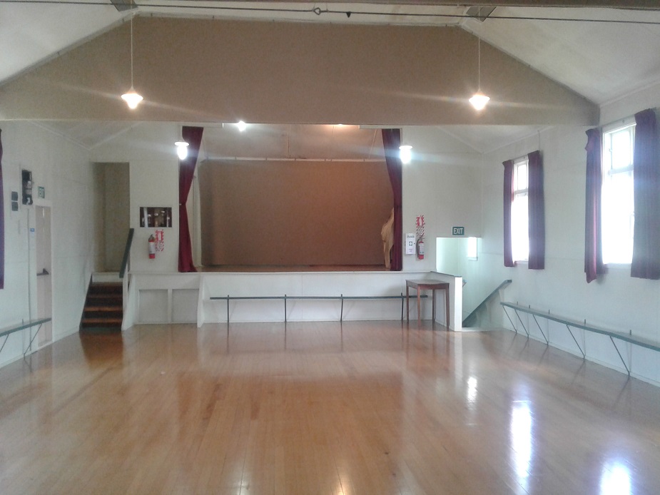 Plain white hall with polished timber floor, raised platform with dark curtains, and strip lighting