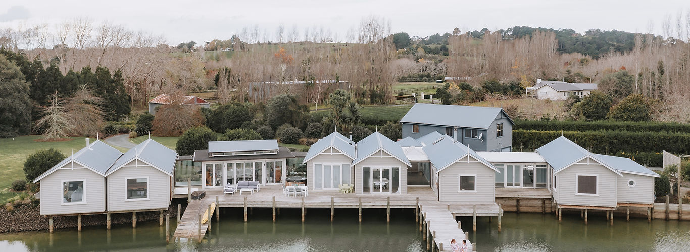Aerial view of a row of white weatherboard boathouses on timber piles over a calm lake with a wooden jetty