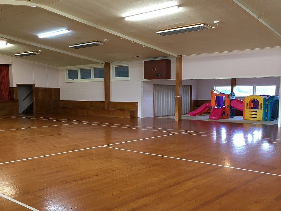 Plain hall interior with timber floor, fluorescent lighting, and children's play equipment in corner