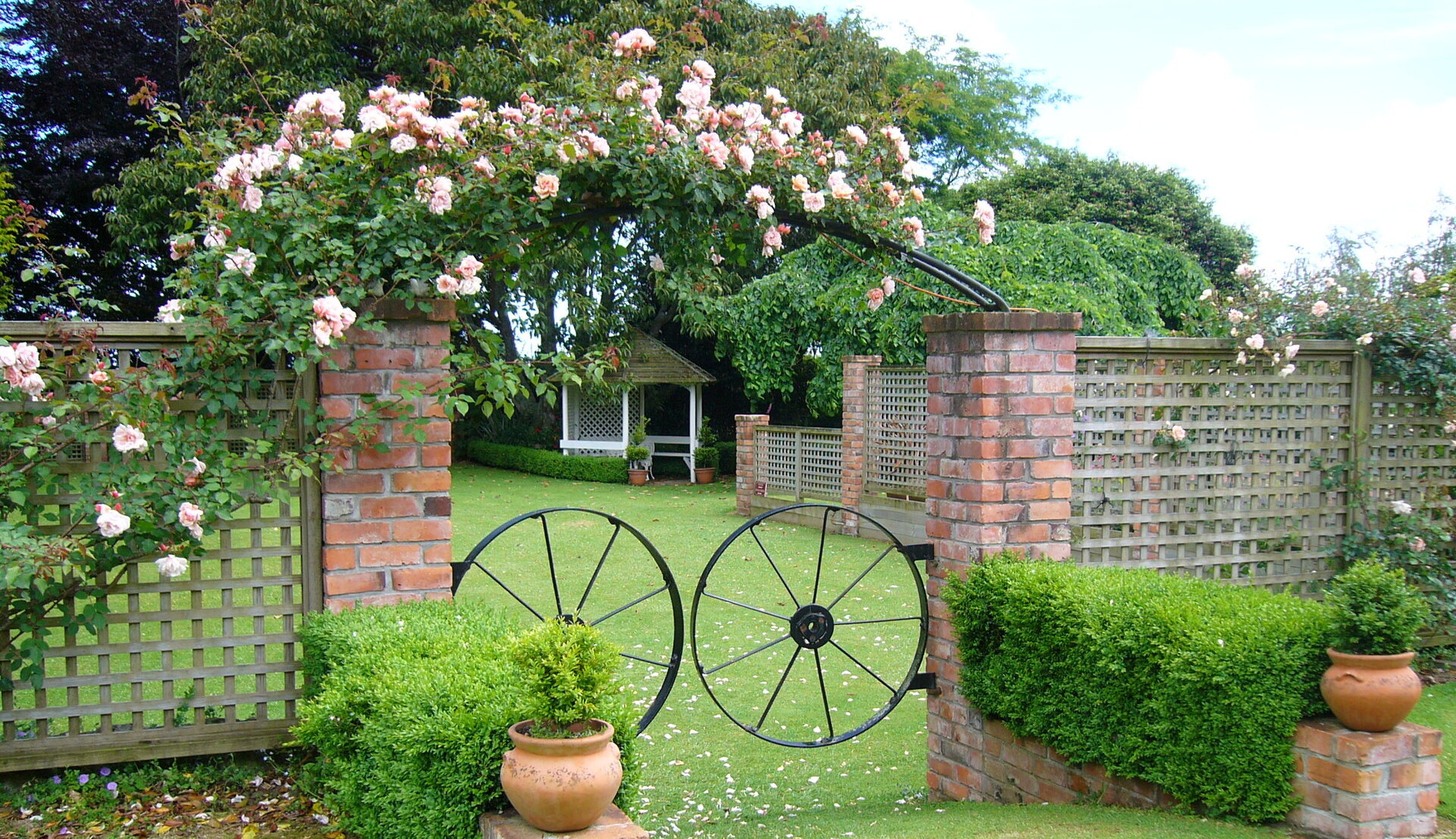 Brick garden gateway with climbing roses and black iron wagon wheels, gazebo beyond on lawn