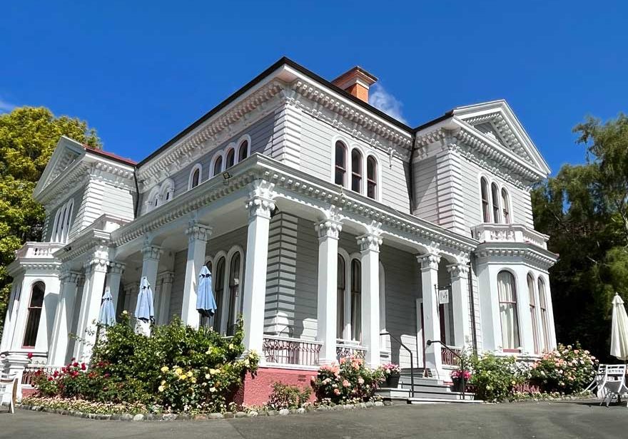 White two-storey Victorian villa with ornate columns, bay windows, and flowering garden beds