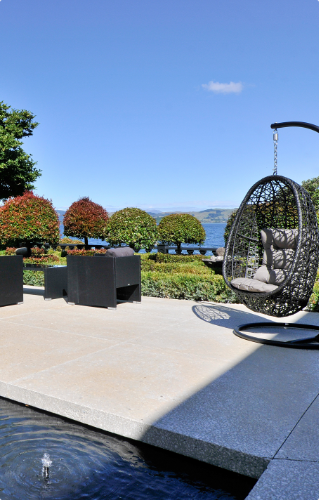 Stone-tiled terrace with suspended egg chair, dark cube seating, and lake and hills view under blue sky