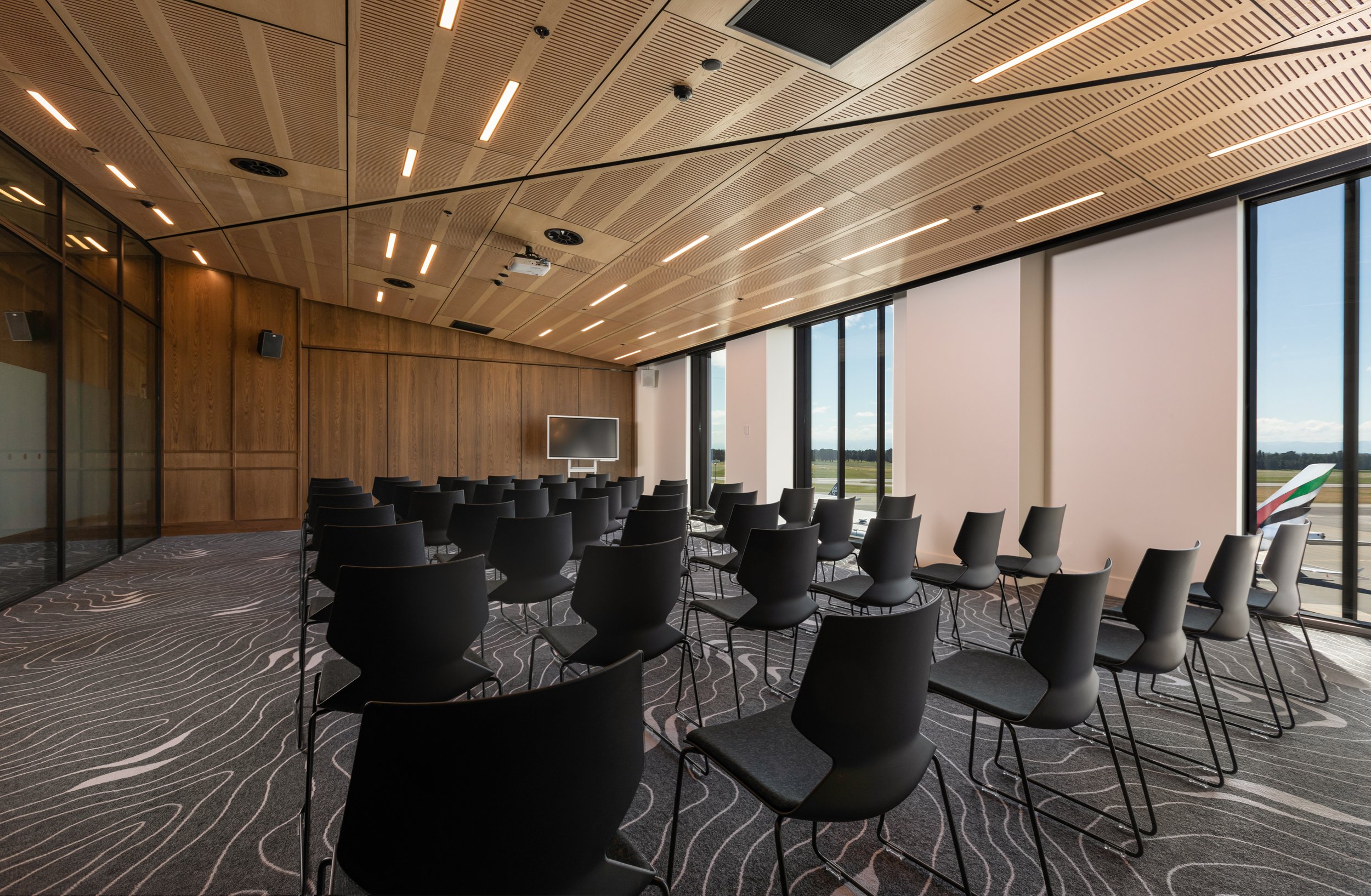 Timber-lined conference room with rows of black chairs, flat screen and large windows on one side