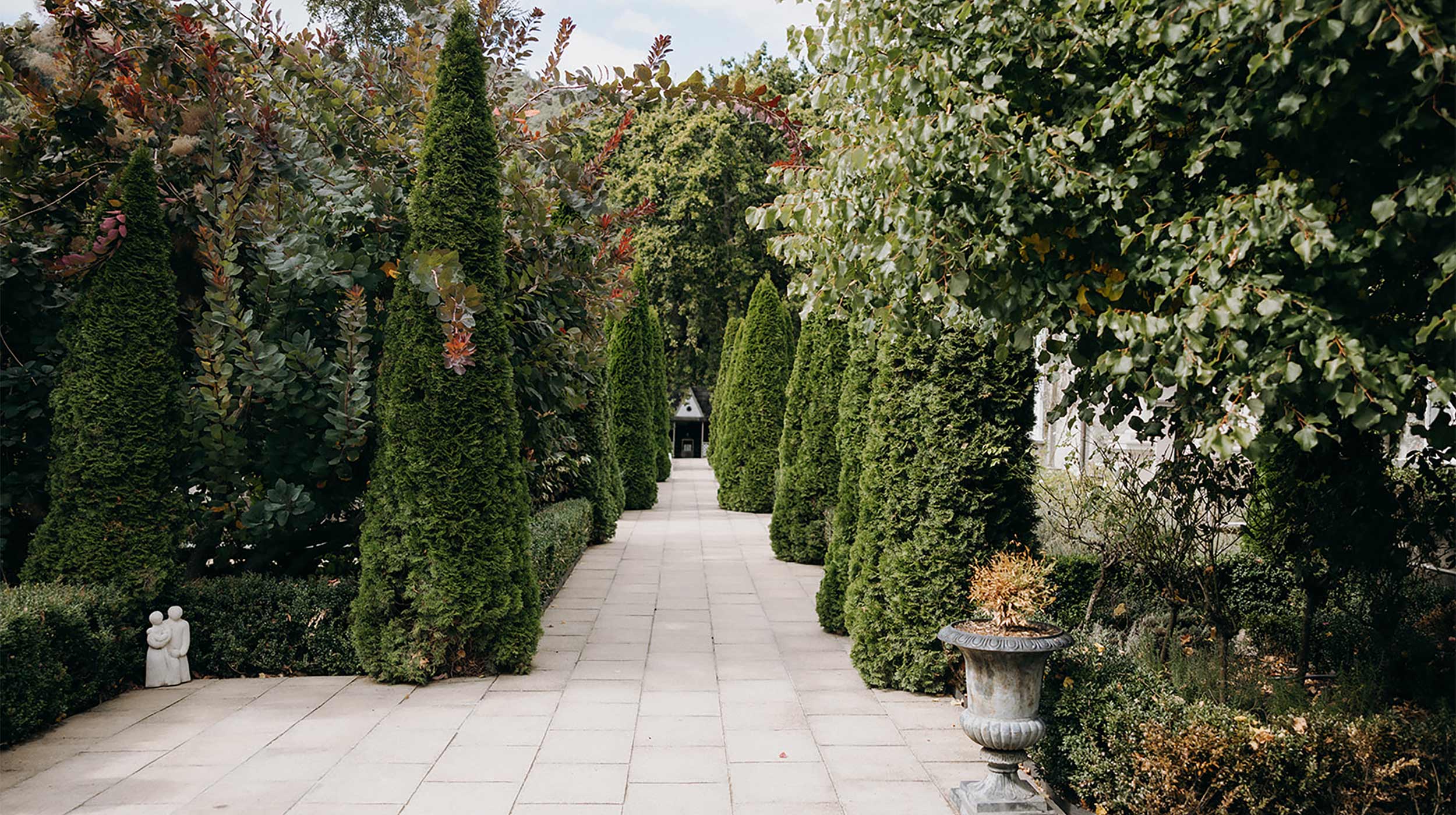 Stone-paved garden path flanked by tall columnar conifers and ornamental planting with an urn