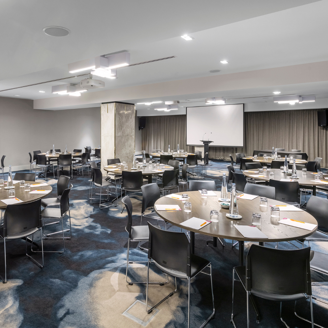 Hotel conference room with round tables, chairs, projector screen, and patterned carpet