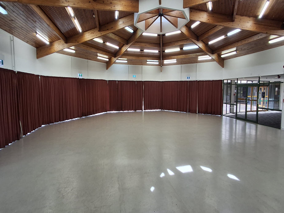 Circular hall with timber beamed roof, concrete floor and red curtains drawn around perimeter